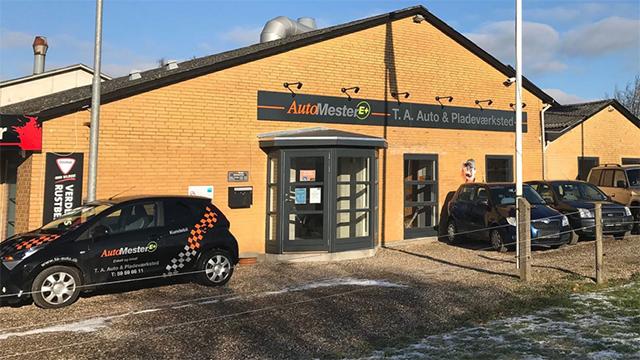Bilforhandler med flere biler parkeret foran en gul murstensbygning med skilt og logo.