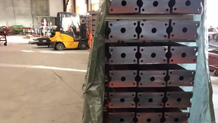 Stabel af metalplader med huller, opbevaret i en lagerhal med en gaffeltruck i baggrunden.