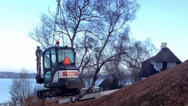 En gravemaskine hæver en træstamme ved en sø, omgivet af en landskab med huse og træer.