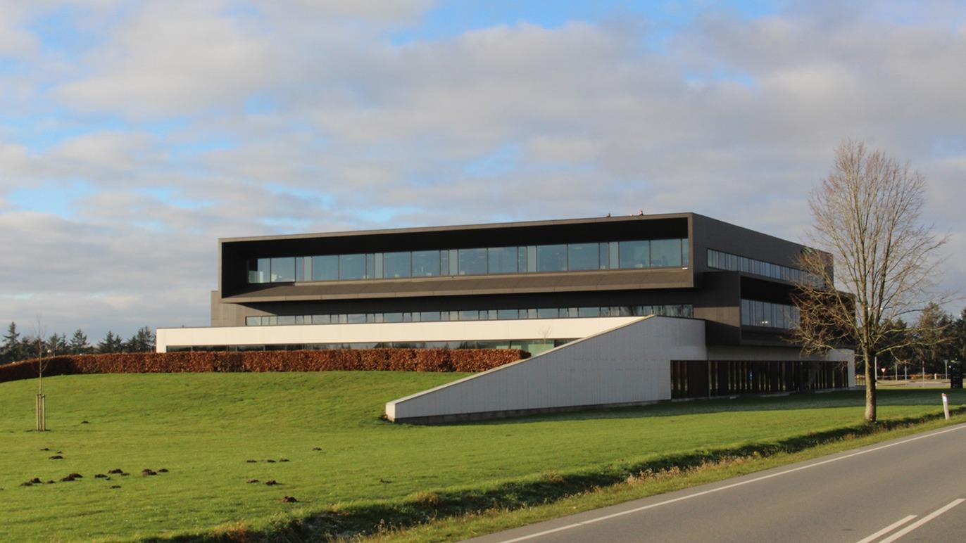 Moderne bygning med store vinduer og skrå tag, beliggende ved en vej og omgivet af grønne områder.
