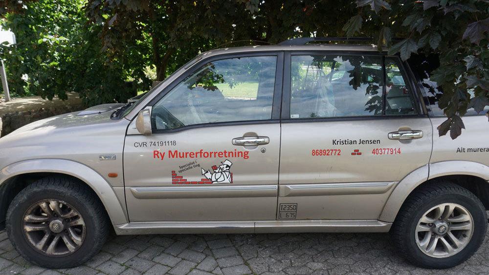 En sølvfarvet bil med reklame for Ry Møbeltransport står parkeret under et træ.