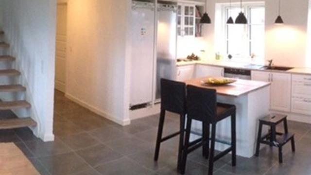 Moderne køkken med en ø, barstole, og trappe til anden sal, belyst af pendellamper.