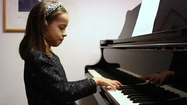 En lille pige spiller klaver i et musiklokale, iført en glitrende bluse og hårbånd.
