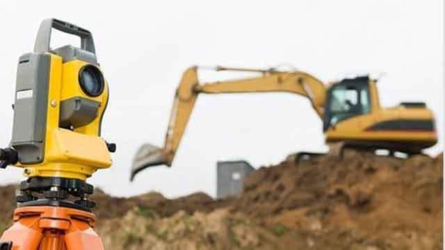 En totalstation i forgrunden med en gravemaskine, der arbejder på en byggeplads i baggrunden.