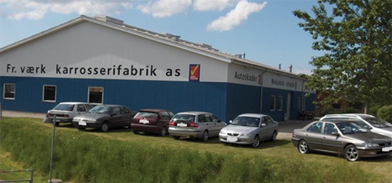 En bilfabrik med flere parkerede biler foran bygningen og en klar blå himmel i baggrunden.