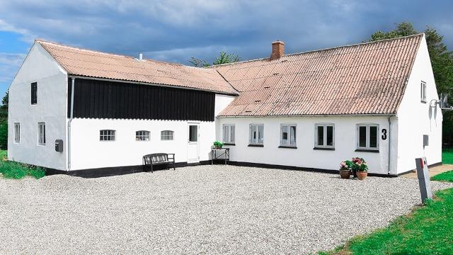 Et hvidt hus med grå grusbelægning foran, omgivet af grønne områder og en blå himmel.