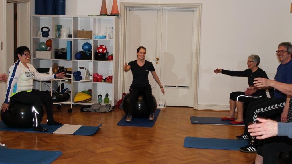 Gruppe af mennesker deltager i en fitnessklasse med træningsbolde og måtter i et lyst lokale.