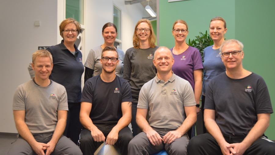 Gruppebillede af ni professionelle terapeuter i træningstøj, smilende og samlet i et lyst lokale.