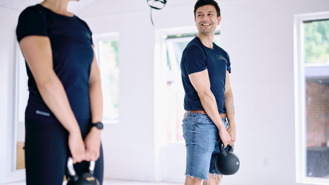 To personer træner med kettlebells i et lyst rum med vinduer. En mand smiler, mens han holder sin kettlebell.