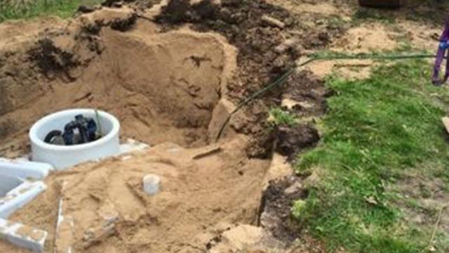 Gravested med en åben grøft, hvor en septiktank er synlig, omgivet af sand og jord.