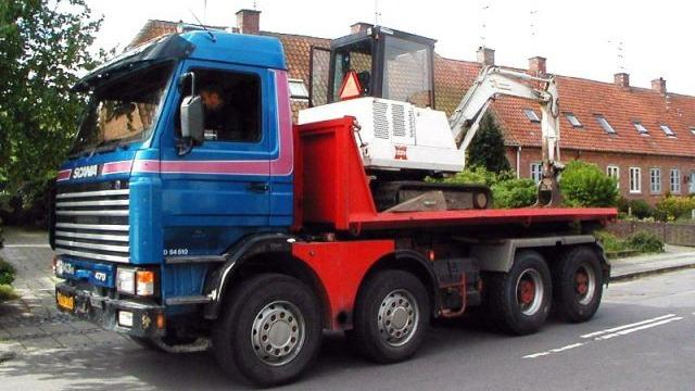 En lastbil med en rød og blå farve, der transporterer en lille gravemaskine på en trailer.