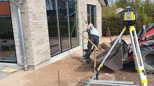 En arbejder graver i sandet ved en bygning, mens en laserafstandsmåler står på stativ i forgrunden.