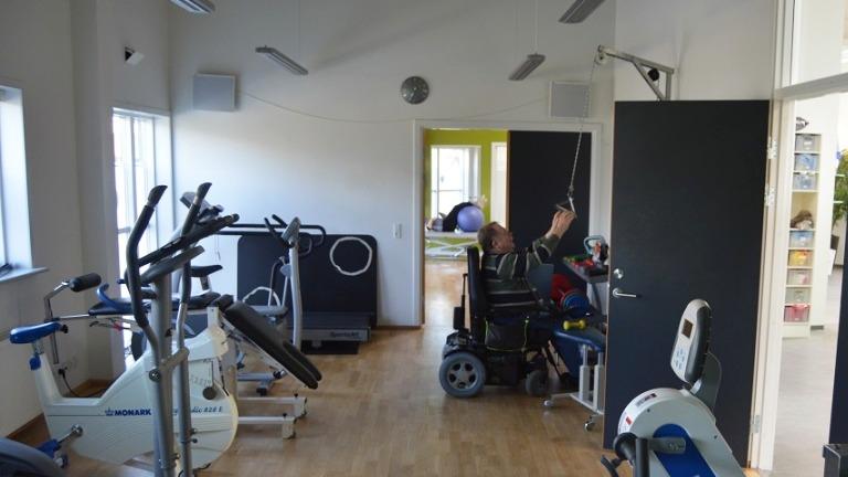 Træningslokale med fitnessudstyr og en person i kørestol, der bruger en træningsmaskine.