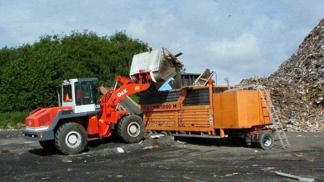 En gummiged læsser affald ind i en stor container på en genbrugsplads med affaldsbunker i baggrunden.