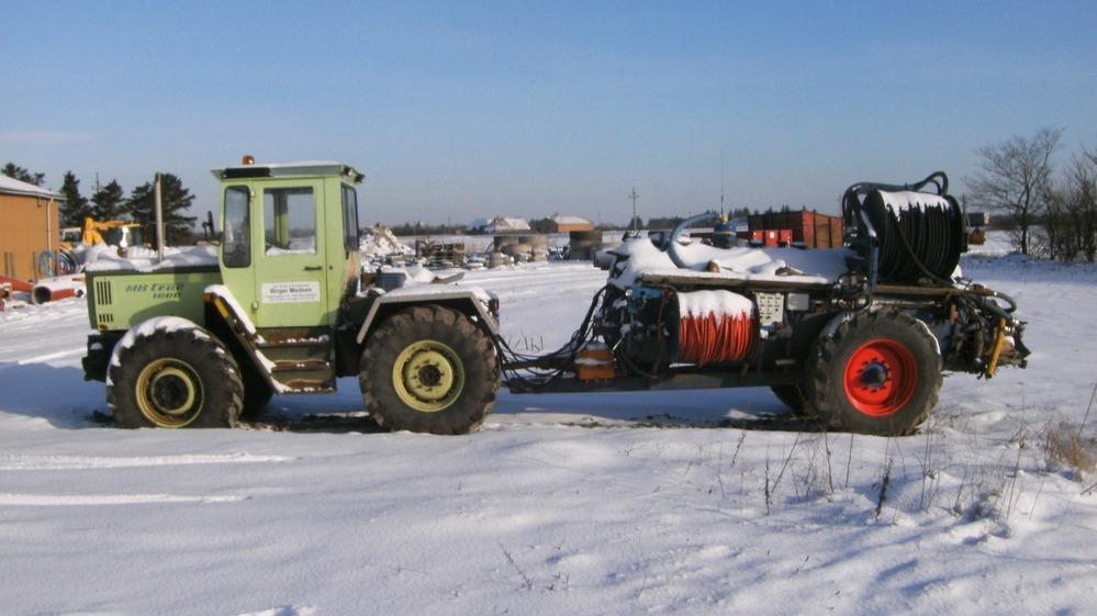 En grøn traktor parkeret i sneen med en stor maskine bagpå, omgivet af en vinterlandskab.