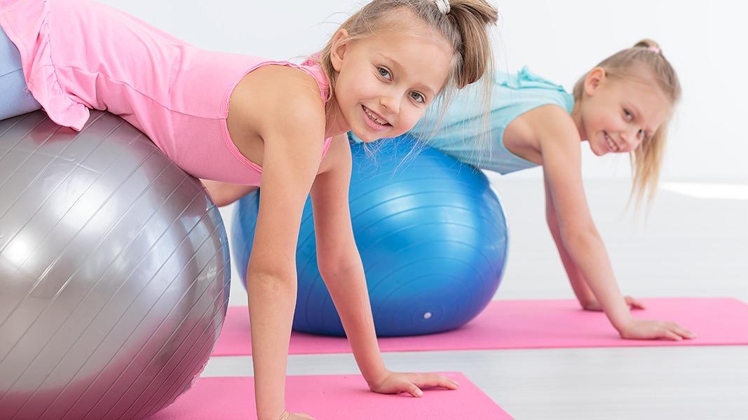 To piger træner på fitnessbolde i et lyst rum med træningsmåtter.