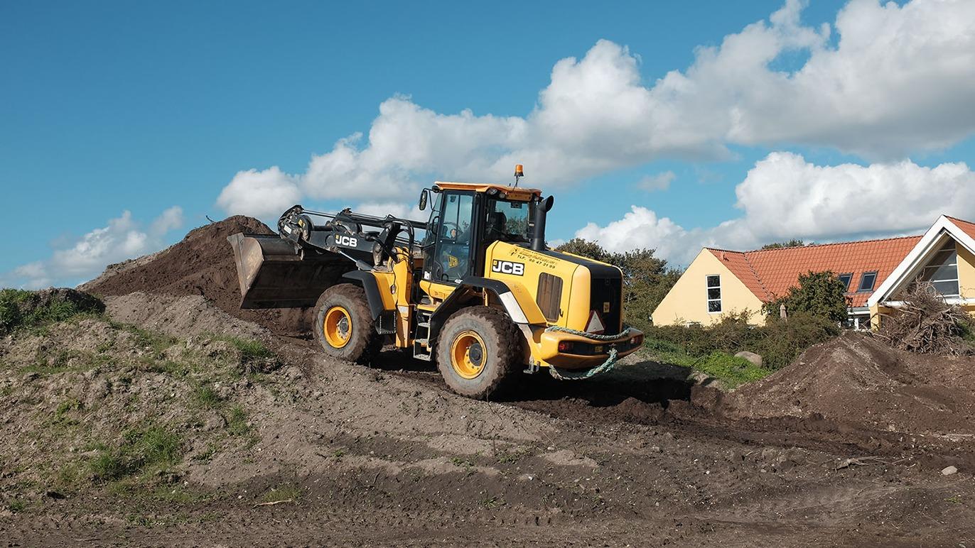 En gult bulldozer arbejder på en byggeplads med en stor bunke jord og en bolig i baggrunden.