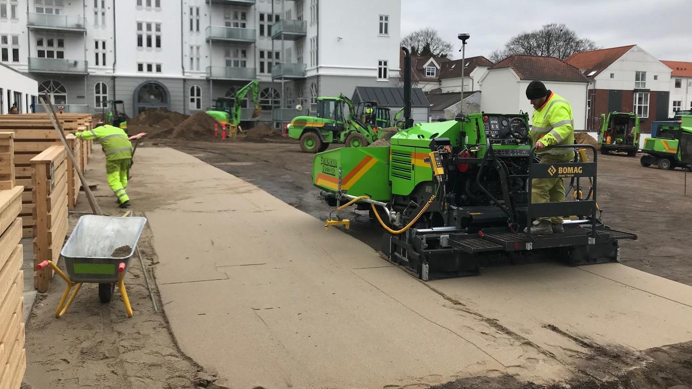 Byggearbejde med maskiner og arbejdere, der lægger beton på en byggeplads med træstakke i baggrunden.