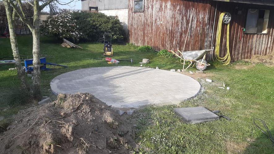 En rund betonplade i en have med græs, omgivet af værktøj og en bygning i baggrunden.