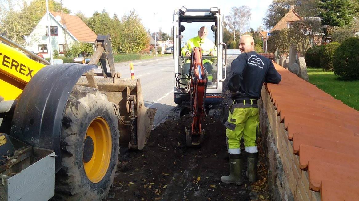 To personer arbejder med gravemaskiner ved en vej, mens de graver i jorden nær en mur.