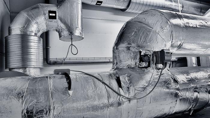 Isolerede rør og ventilationskanaler i en industriel bygning med metaloverflade.
