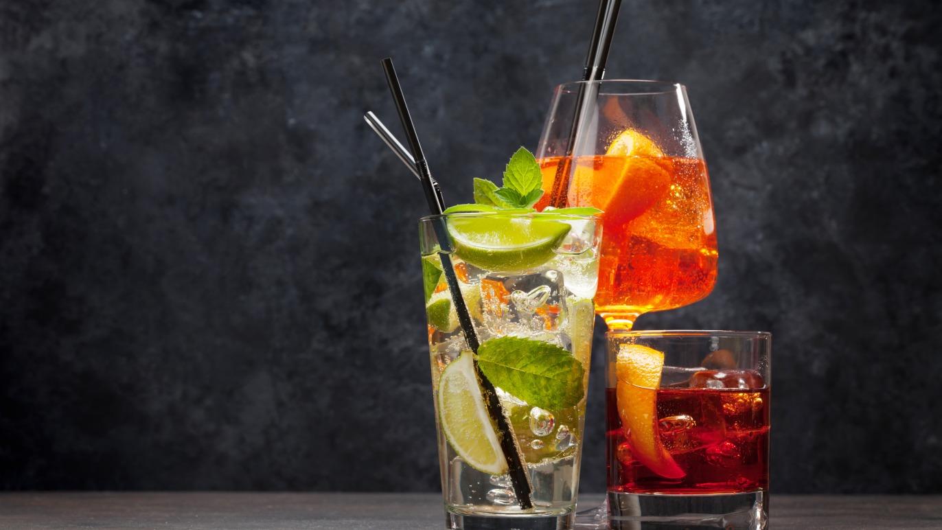 Tre farverige cocktails med lime, mynte og appelsin, serveret i glas med sugerør på en mørk baggrund.