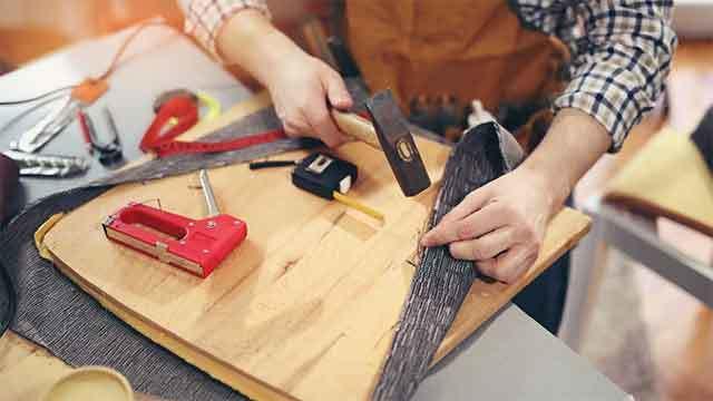 En håndværker arbejder med værktøj på et træbord, mens han fastgør materialer med en hammer og en hæftemaskine.