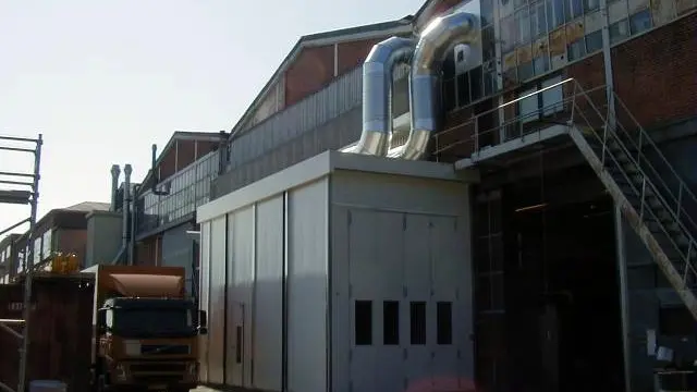 Fabriksbygning med metalrør og lastbil parkeret foran indgangen. Solskin over bygningen.