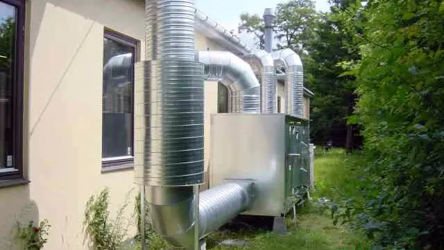 Et hus med metalrør og ventilationssystem placeret ved siden af en have med højt græs.