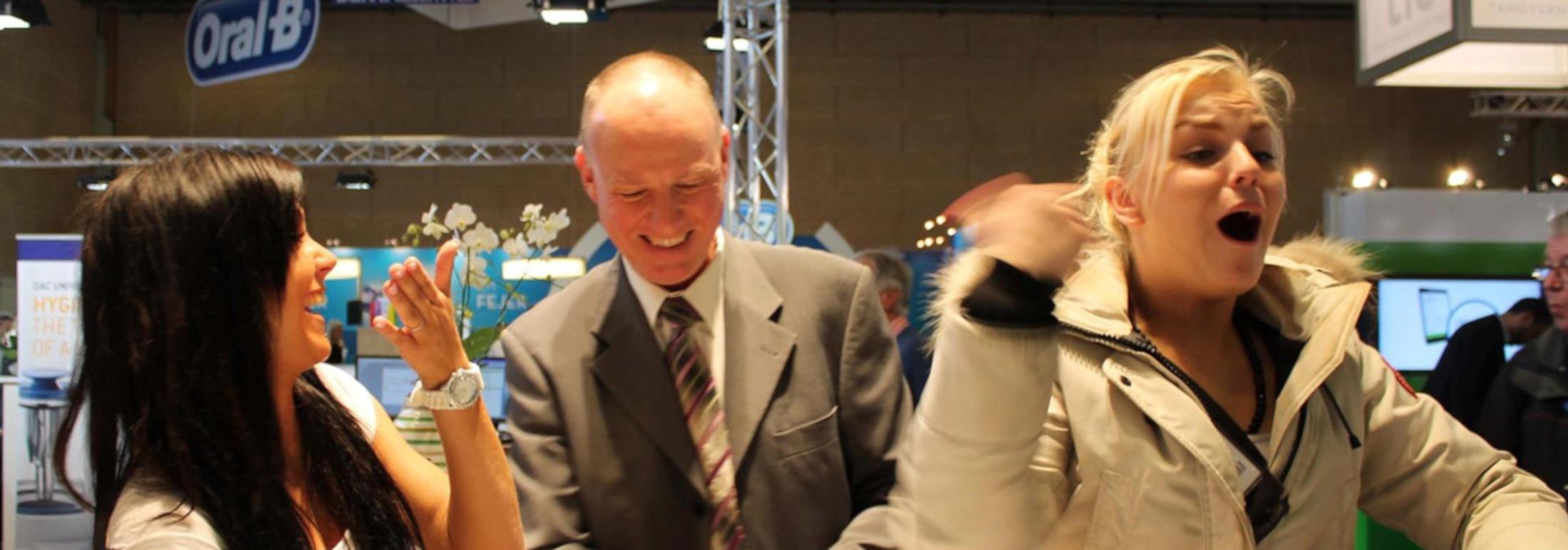 To personer, der griner og interagerer med en ældre mand på en messe, med udstyr i baggrunden.