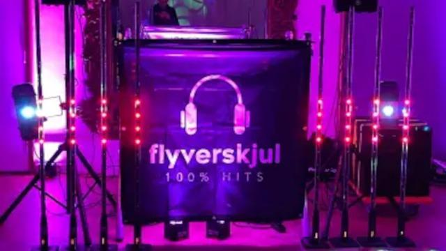 En DJ-bås med lys og højttalere, der præsenterer "flyverskjul 100% hits" i et festligt miljø.