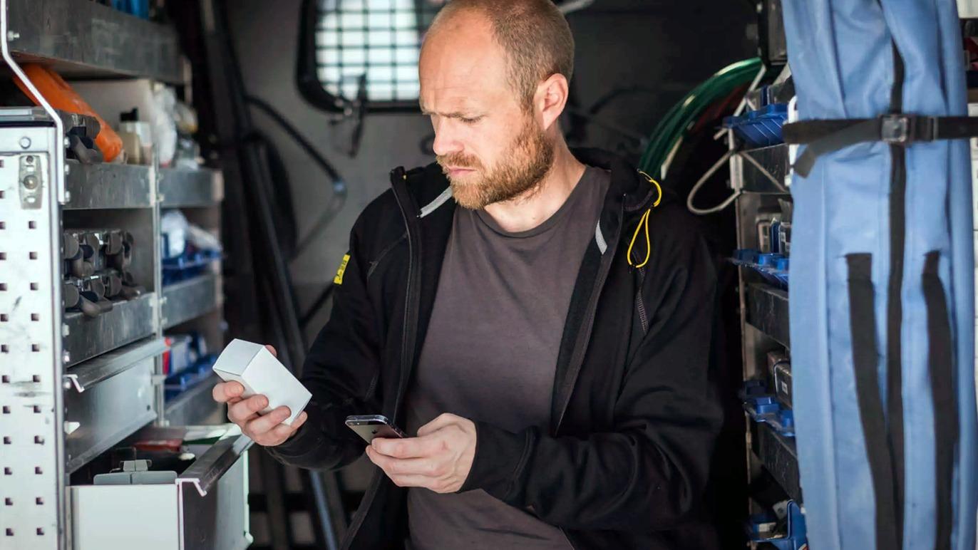 En mand med skæg står i en varevogn og kigger på en enhed, mens han holder en smartphone i hånden.