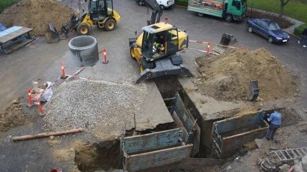 Billede af en byggeplads med gravemaskiner, grus og en åben grav til rørarbejde.