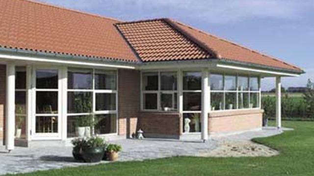Moderne hus med rødt tag og store vinduer, beliggende i en grøn have med flisebelægning.
