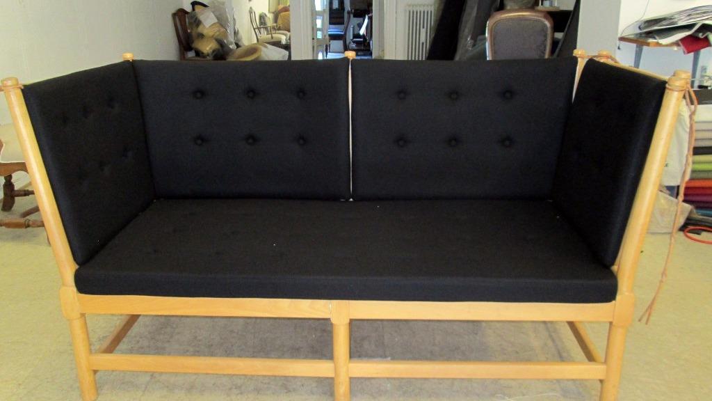 Sort sofa med træramme og knapper, placeret i et lyst rum med andre møbler i baggrunden.