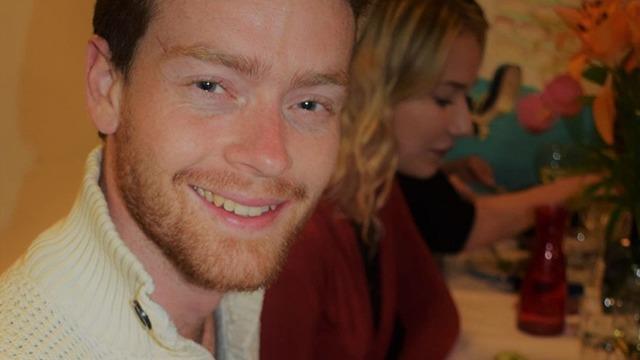 En smilende mand med rødt skæg sidder ved et bord, mens en kvinde arbejder i baggrunden.