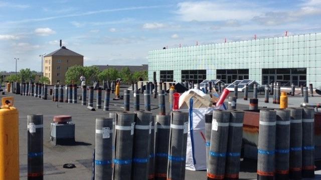 Lagerområde med mange rør og containere, bygning i baggrunden under en blå himmel.