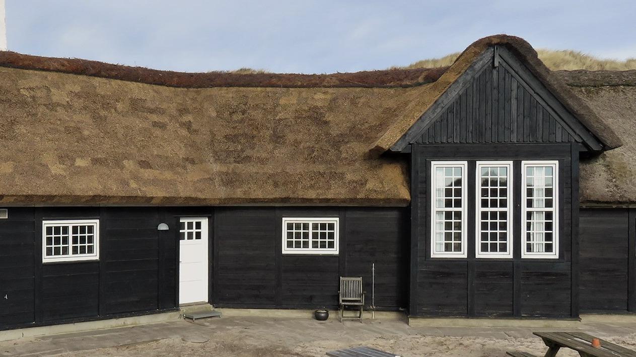 Et sort hus med stråtag og store vinduer, beliggende i et naturskønt område.
