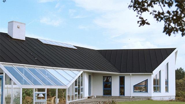 Moderne hvidt hus med sort tag og glasvæg, omgivet af grønne områder og træer.