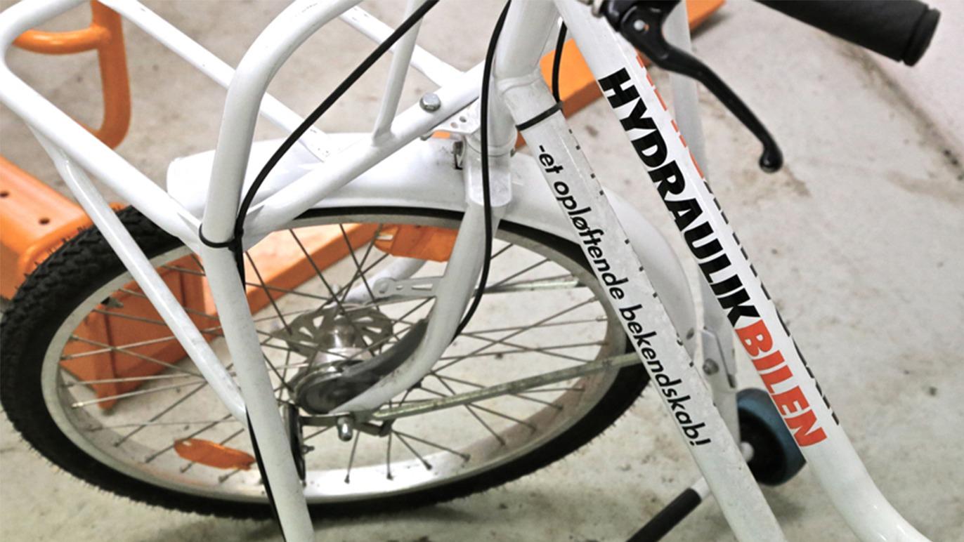 Billede af en hvid cykel med teksten "HYDRAULIKBIEN" på stellet og en bagagebærer i orange.