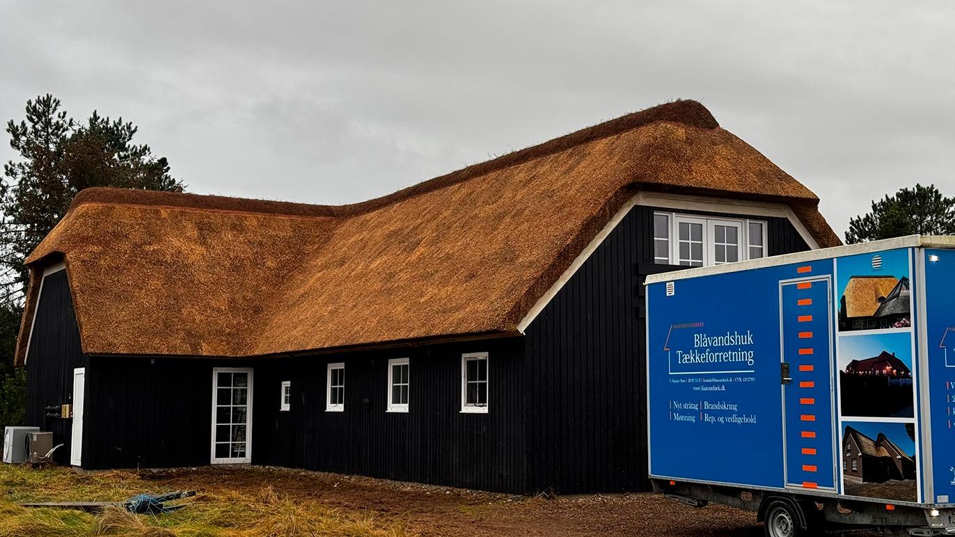 Et sort hus med stråtag og en blå lastbil parkeret foran. Huset har store vinduer og ligger i et naturskønt område.