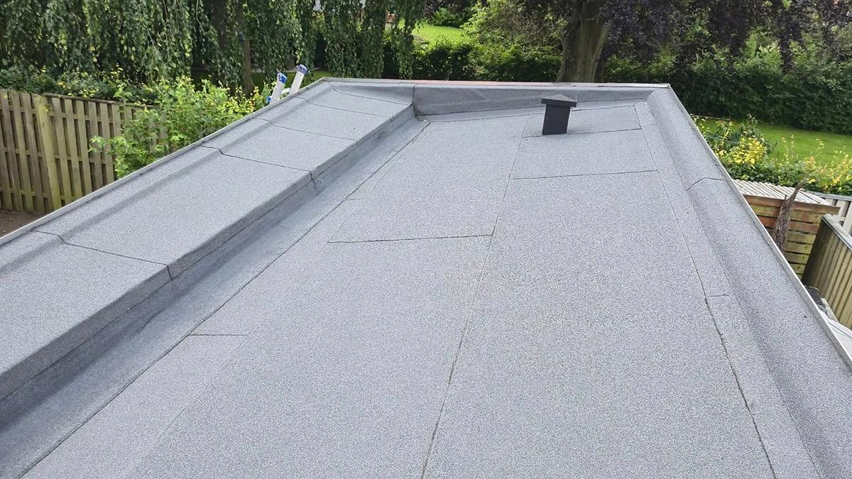 Fladt tag med grå belægning og ventilationsrør, omgivet af grønne træer og en have.