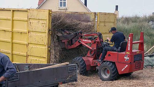 En landmand bruger en rød traktor til at læsse halm i en gul container ved en gård.