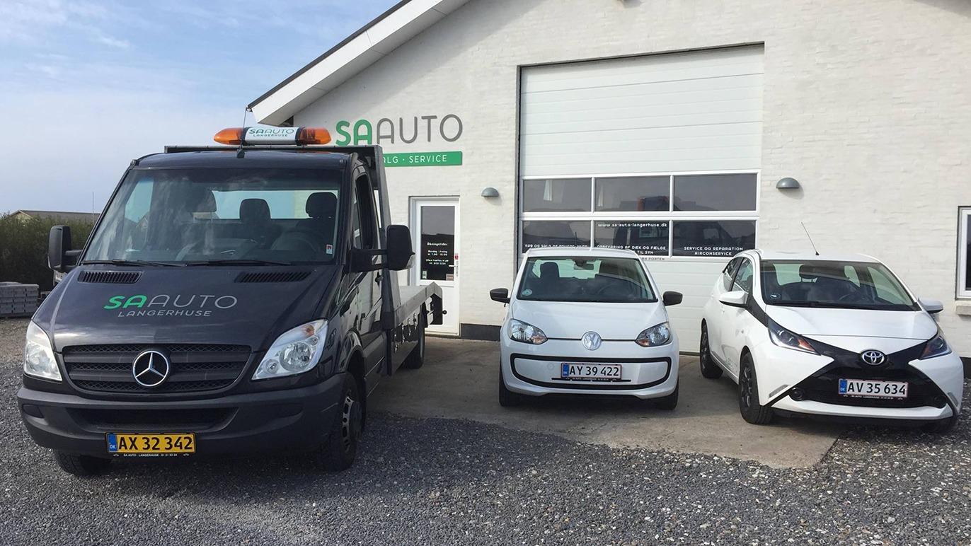 En sort lastbil og to hvide biler parkeret foran en bilforhandler med logoet "SA Auto" på bygningen.