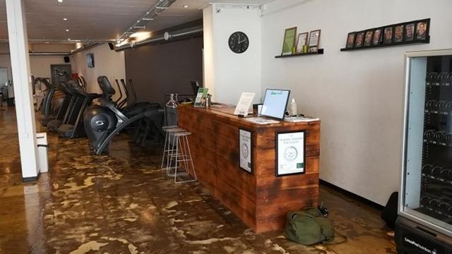 Modtagelsesområde i et fitnesscenter med trædisk, træningsudstyr og en væg med billeder.