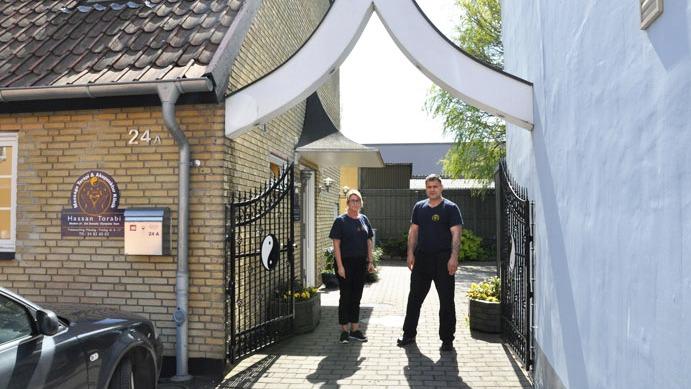 To personer står ved indgangen til en gård med en hvid bue og murstensvægge i sollys.