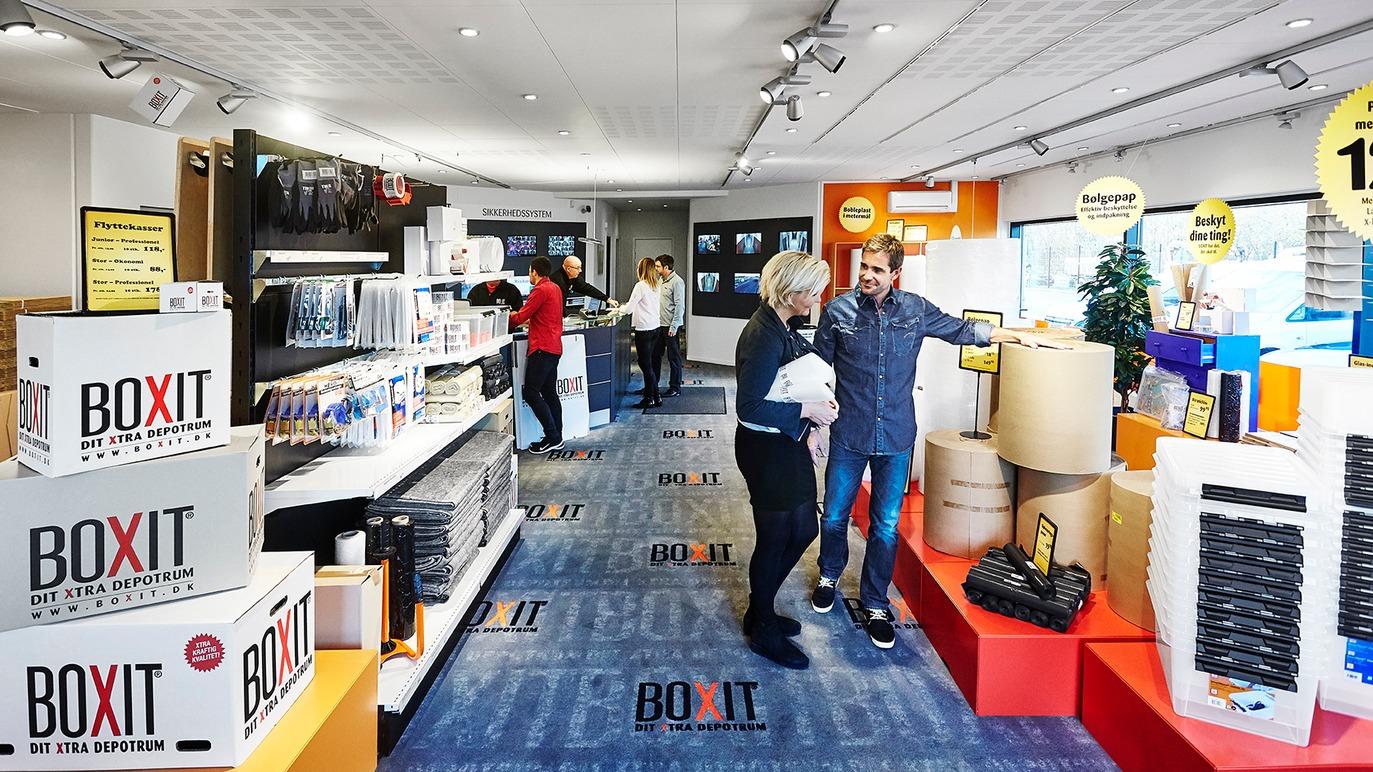 Butik med kunder, der handler, og hylder fyldt med kontorartikler og emballageprodukter.