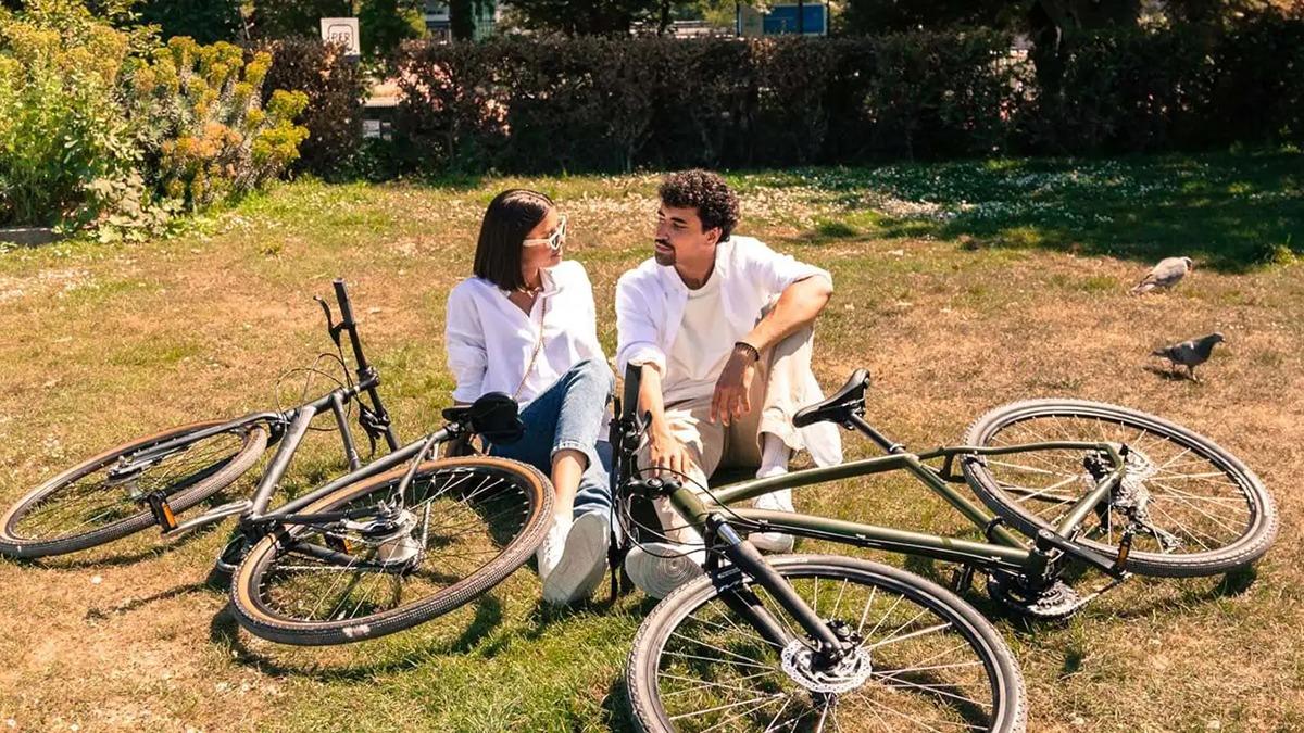 To personer sidder på græsset med cykler omkring dem, mens de snakker og nyder solen.