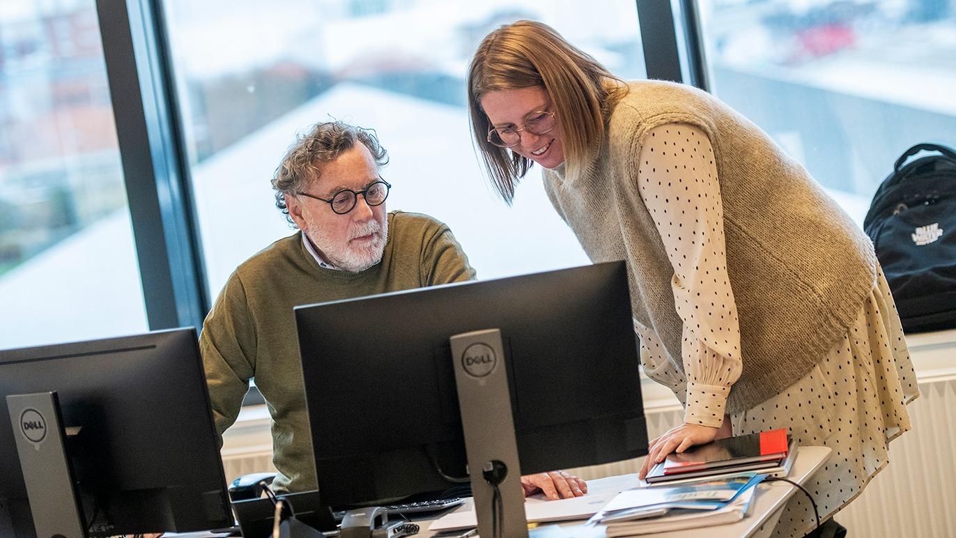 To personer, en mand og en kvinde, arbejder sammen ved et skrivebord med computere i et lyst kontormiljø.