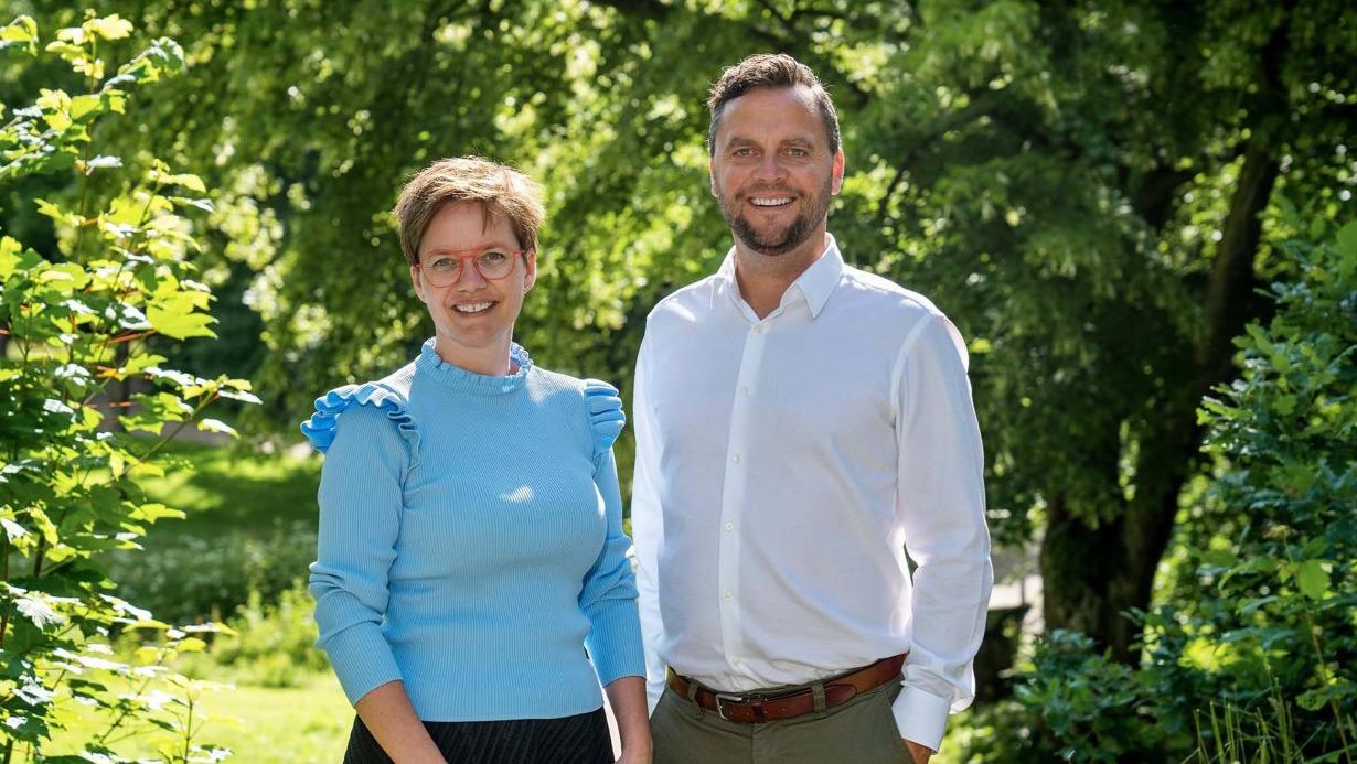 To personer står smilende i en grøn park med træer i baggrunden, iført afslappet tøj.